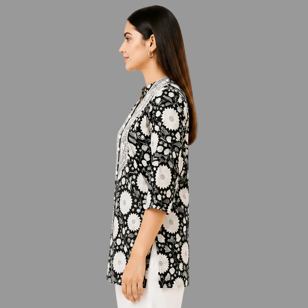 A woman shown in side profile wearing a black tunic-style top with a white floral and circular print, paired with white pants, against a plain white studio background.