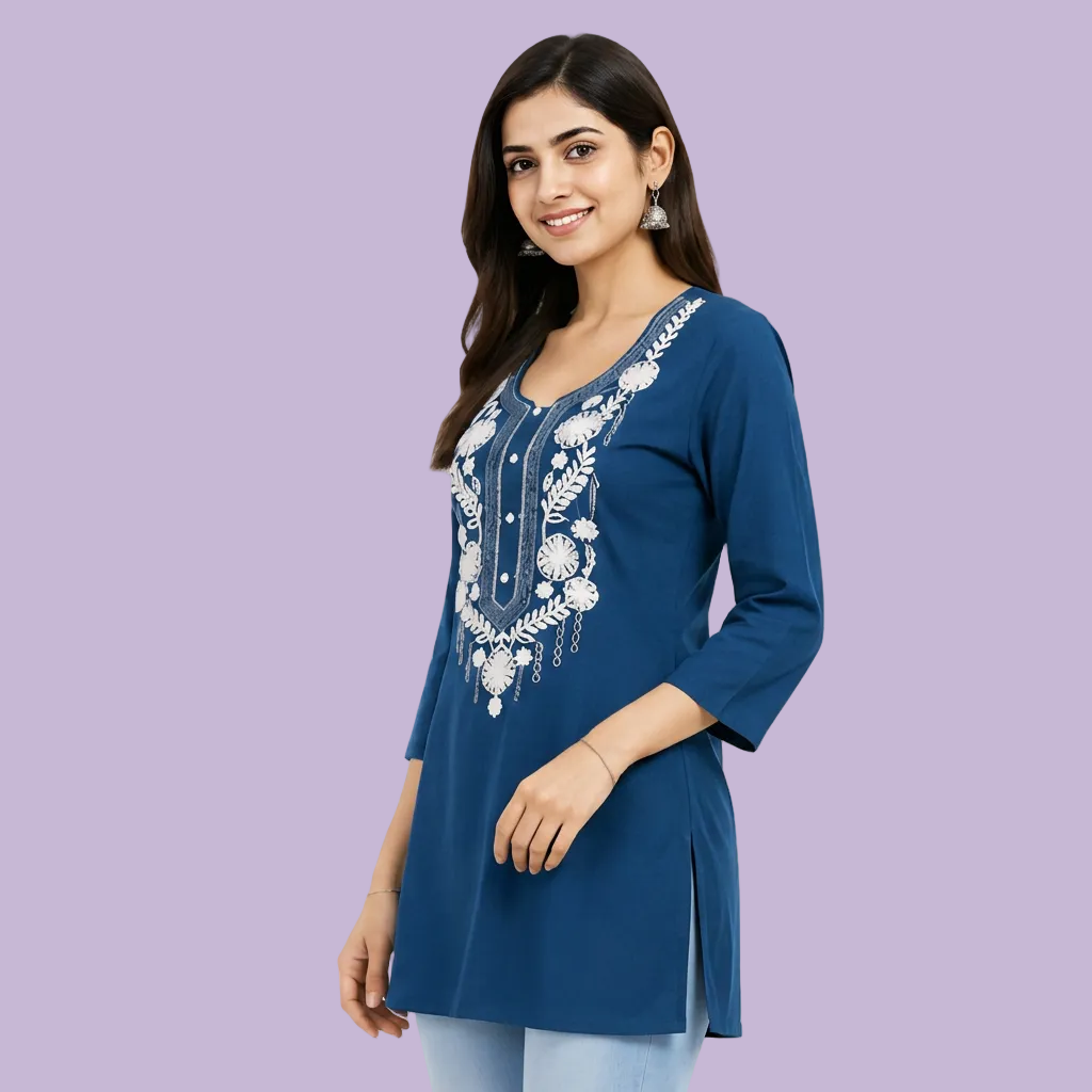 A woman modeling a royal blue tunic top with white floral embroidery on the front, paired with light blue jeans against a plain light gray studio background.