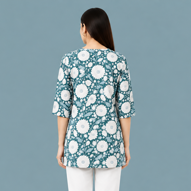 A woman viewed from the back wearing a teal three-quarter sleeve top with a white floral pattern, paired with white pants, against a plain white background.