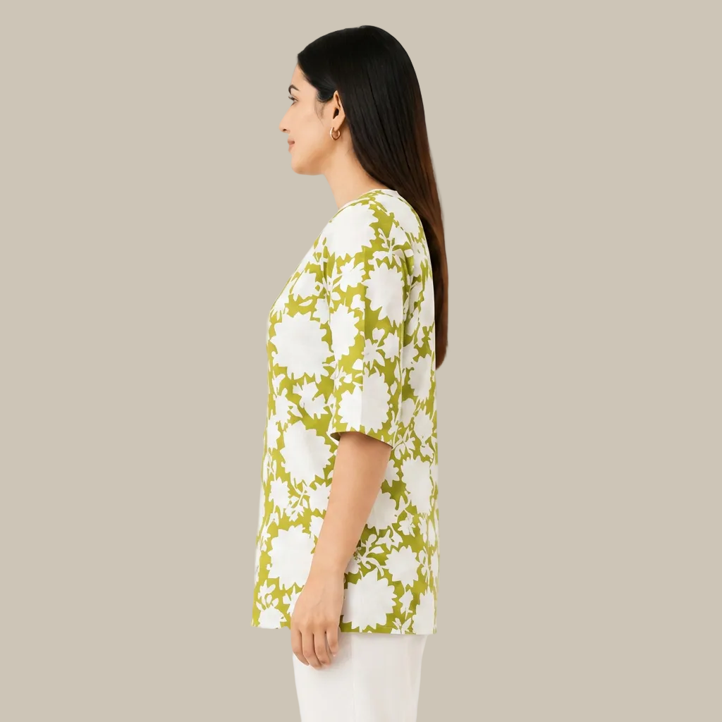 A woman shown in side profile wearing a short-sleeve top with a bold white floral pattern on a lime green background, paired with white pants, against a plain white studio backdrop.
