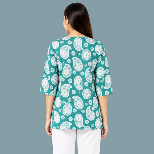 A woman viewed from the back wearing a teal green top with a white paisley pattern and white pants, set against a plain white background.