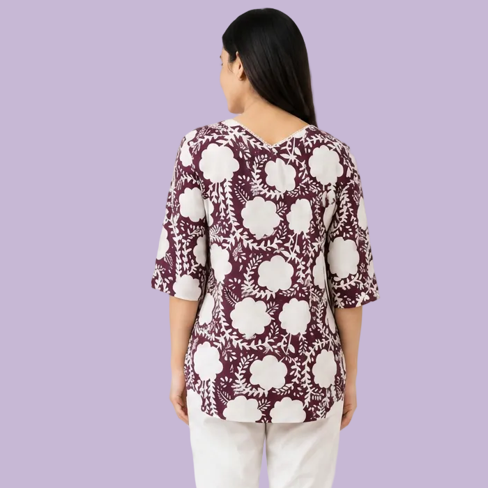 A woman shown from the back wearing a short-sleeve V-neck top with a white floral block print on a deep plum purple background, paired with off-white pants, against a plain white studio background.