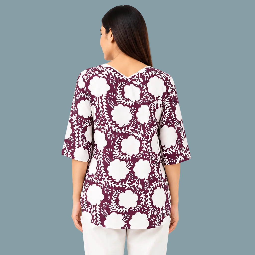 A woman shown from the back wearing a short-sleeve V-neck top with a white floral block print on a deep plum purple background, paired with off-white pants, against a plain white studio background.