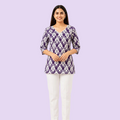 A woman modeling a purple and white patterned tunic top with three-quarter sleeves and a V-neckline, paired with white pants, against a plain white background.