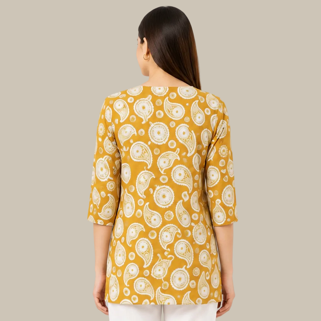 A woman viewed from the back wearing a mustard yellow tunic top with a white paisley pattern, paired with white pants, against a plain white background.