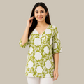 A woman modeling a green and white floral print tunic top with three-quarter sleeves and a V-neckline, paired with white pants against a plain white background.
